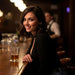 A woman with long brown hair holding a drink, standing in a dimly lit setting with people in the background.
