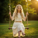 A woman with blonde hair sitting on a swing in a park, wearing a white dress, with sunlight filtering through trees.