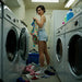 A woman with short brown hair doing laundry in a laundry room, holding a basket of clothes, with a washer and dryer in the background.