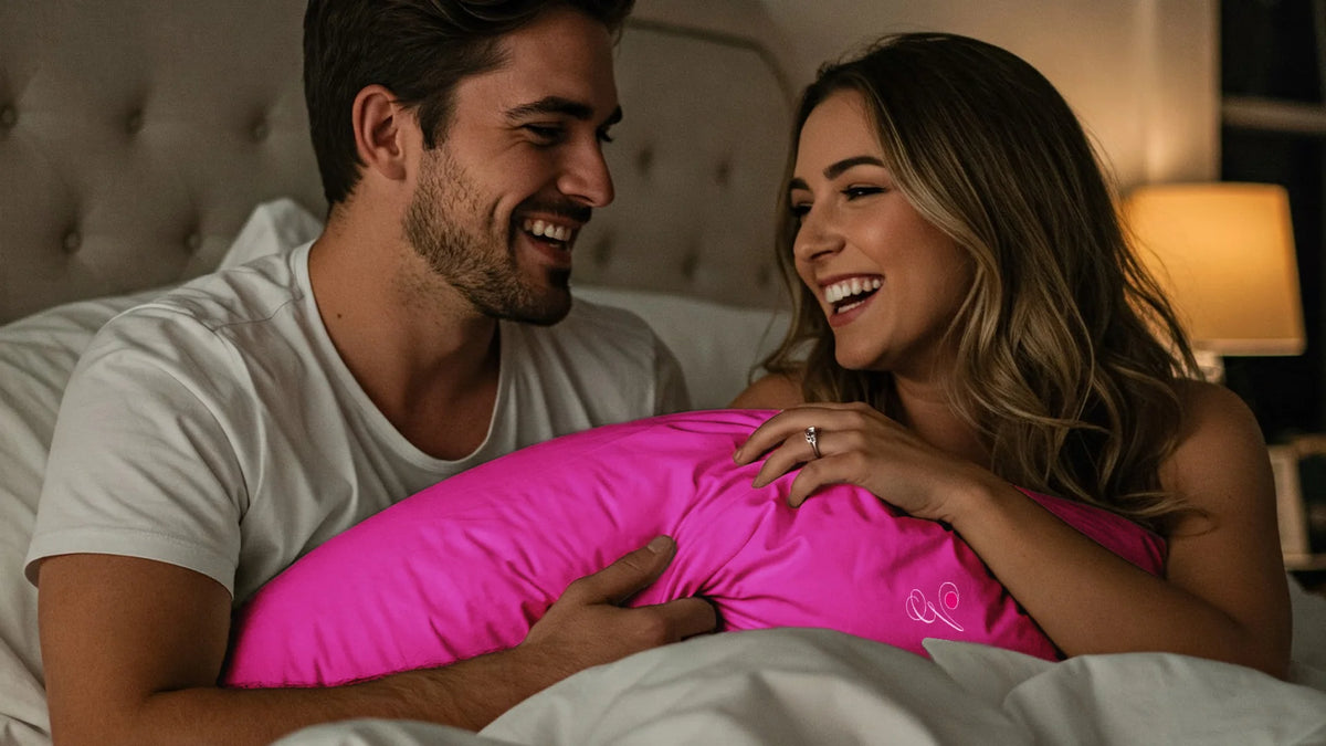 Flirty couple play on a bed with a pink Pillowsforeplay pillow between them